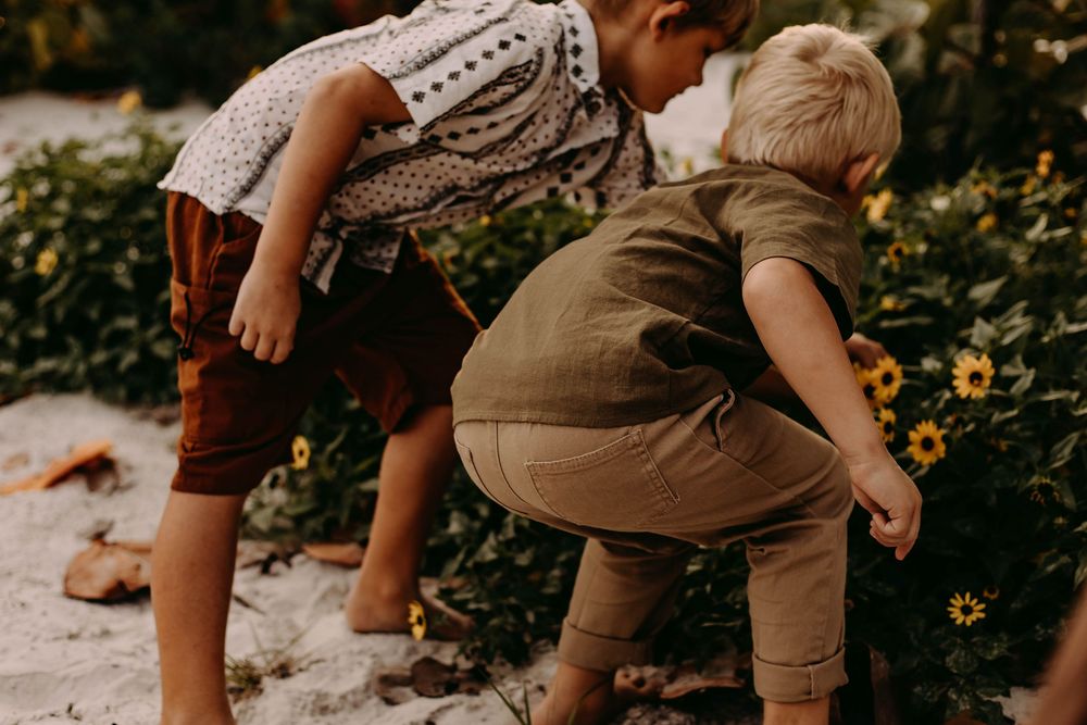 A child in brown overalls explores a garden filled with yellow flowers and green plants on a sunny day.
