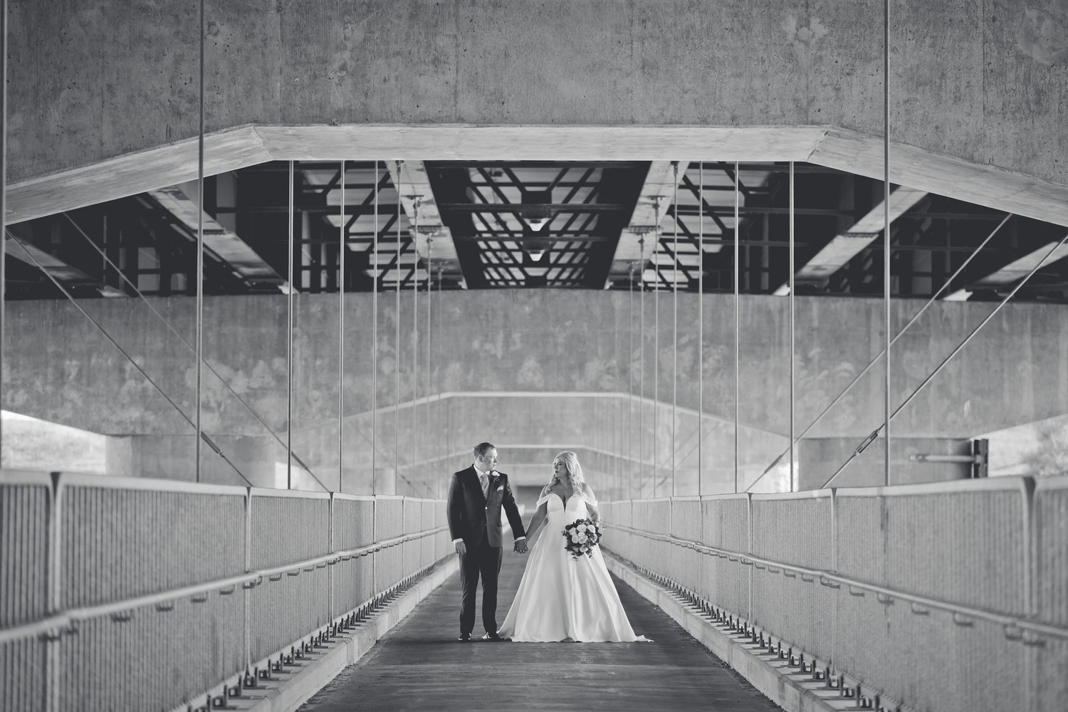 Two people stand at the end of a long, empty concrete hallway with fluorescent lighting overhead.