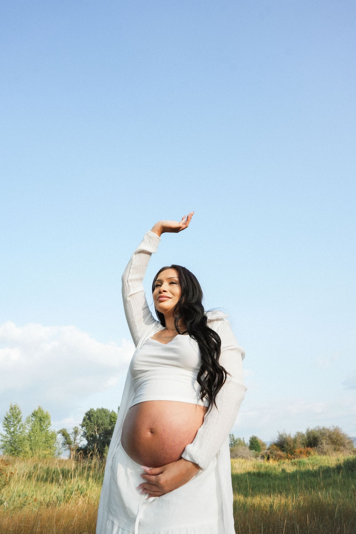 Expectant mother in white dress poses gracefully in a sunny meadow with tall grass and blue sky.