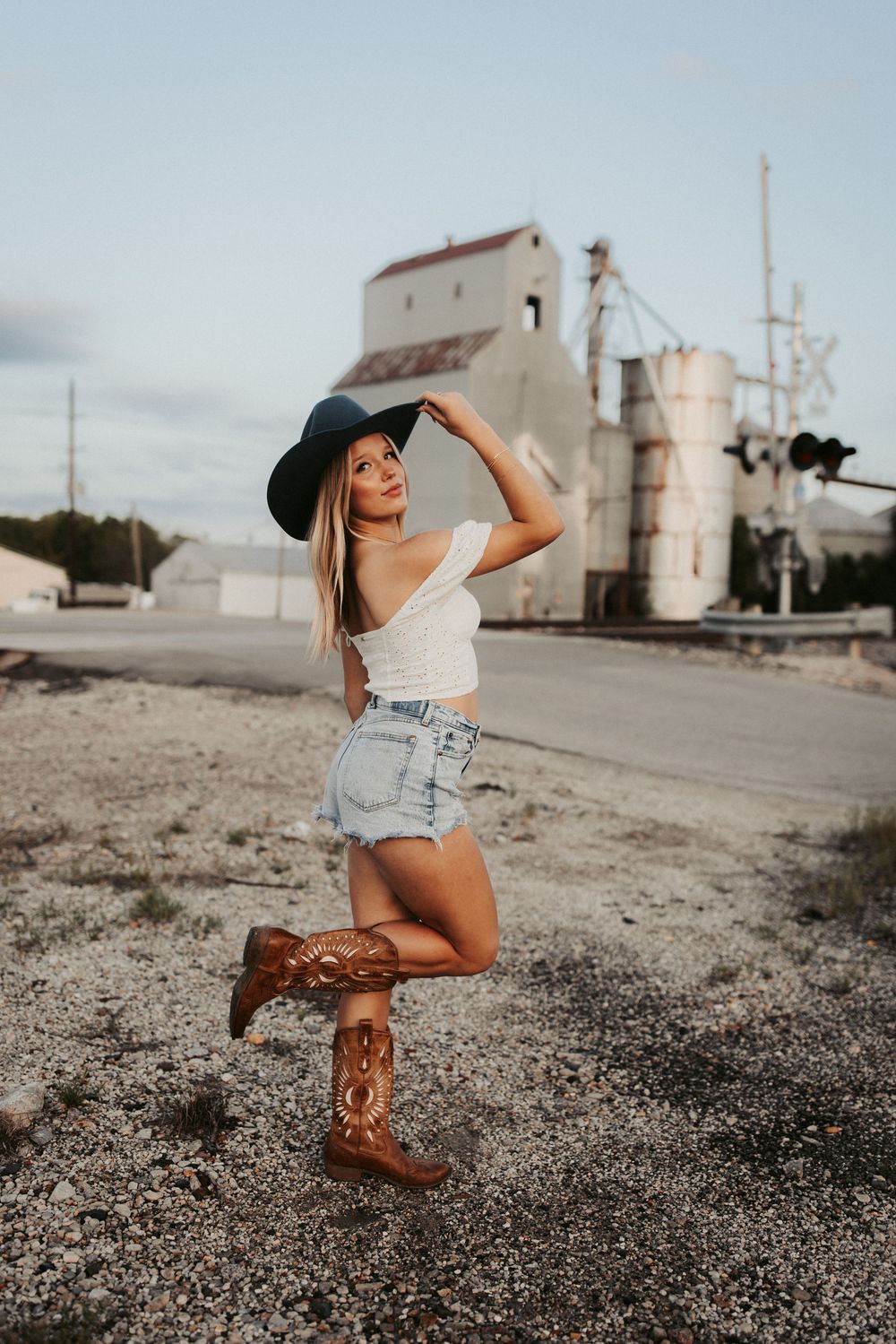 Stylish western outfit with cowboy boots and hat posed in front of industrial grain silo building at sunset.