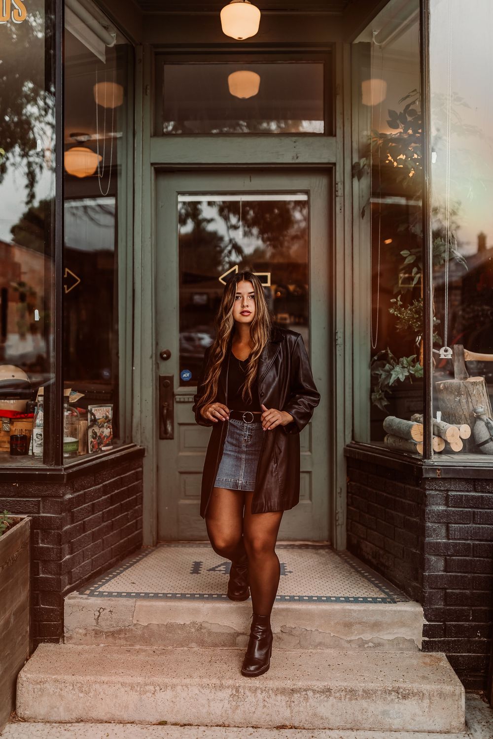 A woman in a leather jacket and skirt stands in front of a storefront with glowing lantern lights at sunset.