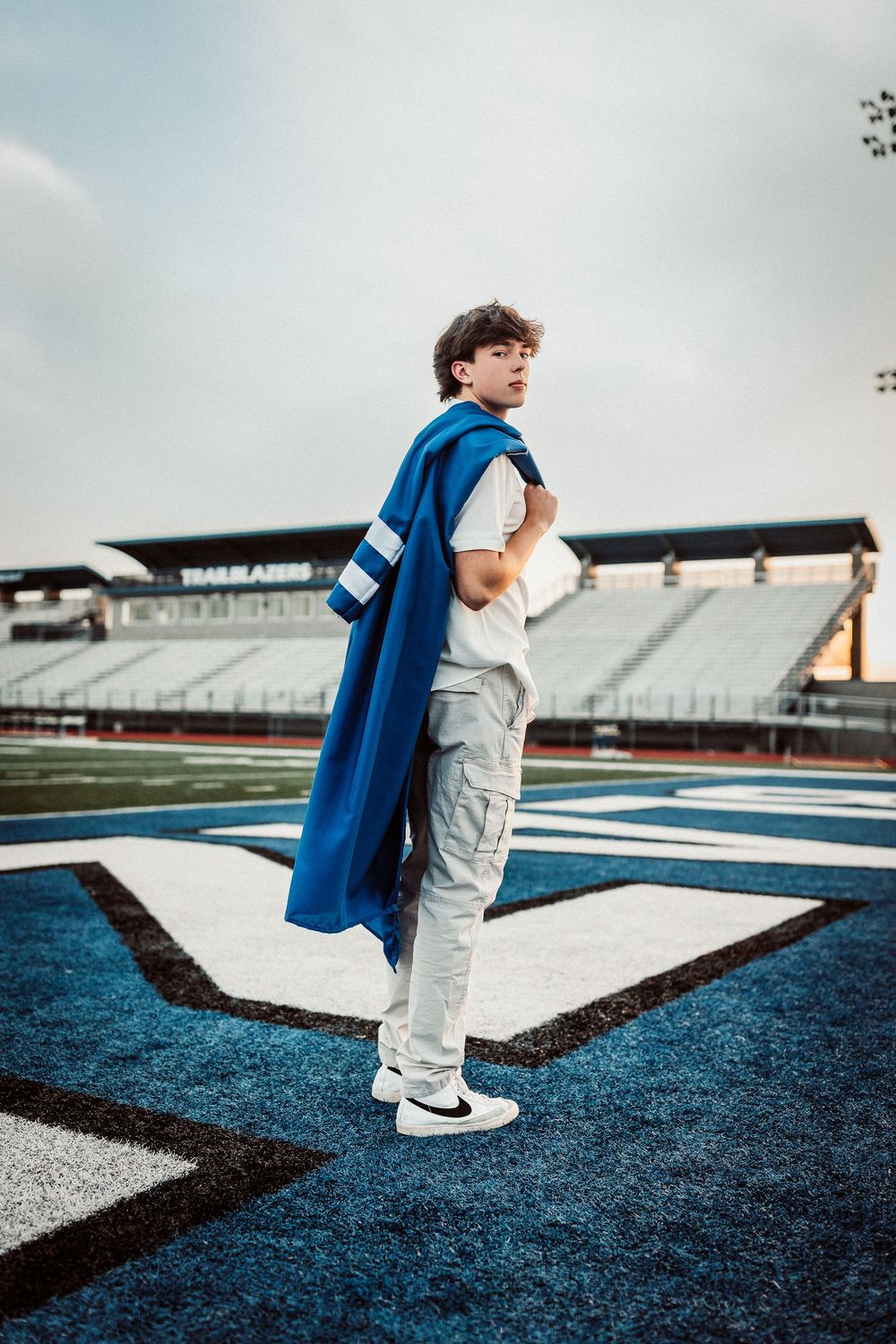 A person wearing a blue graduation gown stands on a football field with stadium bleachers in the background.