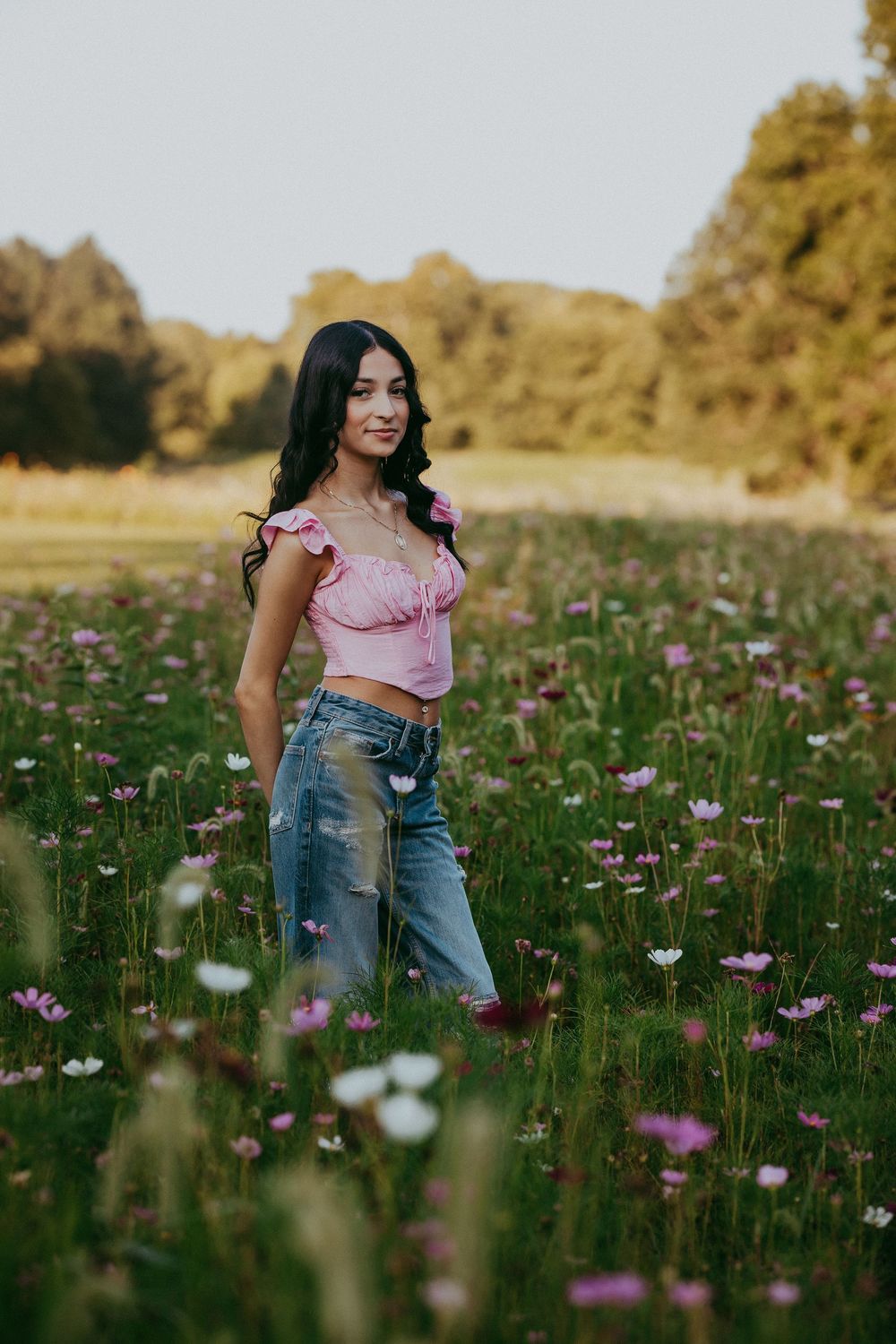 Person in pink top and jeans stands joyfully in a field of colorful wildflowers during golden hour.