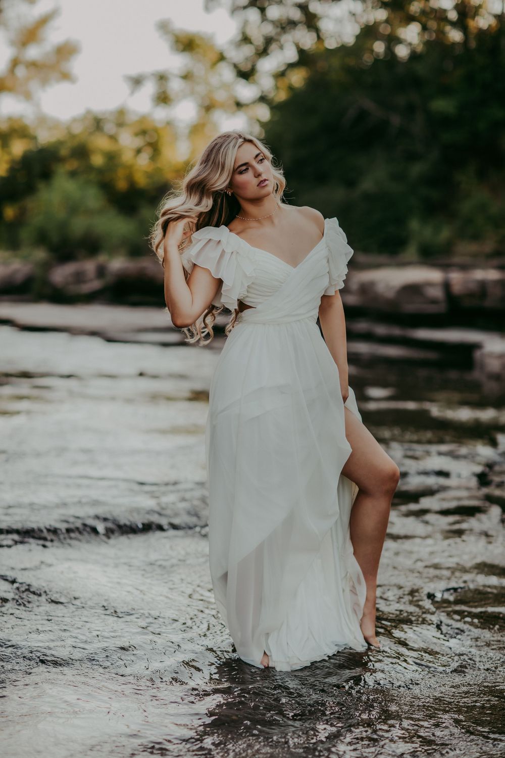 A woman in a flowing white dress poses gracefully on rocks near a serene river during golden hour.