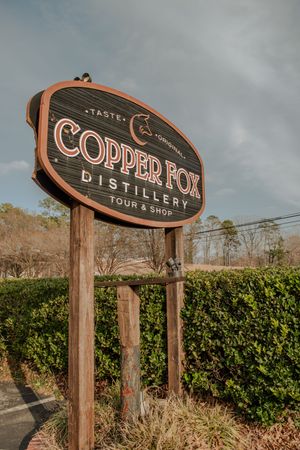 Wooden sign for Copper Fox Distillery mounted on rustic posts against a cloudy sky and green hedges.