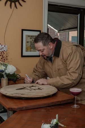 Person in brown jacket writing or signing documents at a wooden table with a cocktail glass.