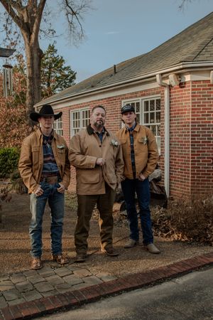 Three people in western-style workwear stand outside a brick building on a paved walkway.