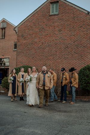 Wedding party in brown attire poses in front of brick building during outdoor celebration.