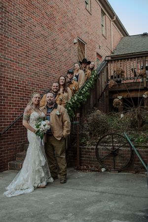 Wedding party poses on brick steps wearing rustic brown attire with bride in lace dress holding white bouquet.