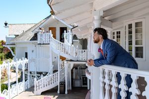 A person in a navy blazer leans on an ornate white balcony railing of a traditional Victorian-style house.