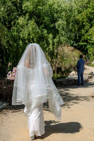 A bride in a flowing white dress and veil walks along a sunlit garden path surrounded by lush greenery.