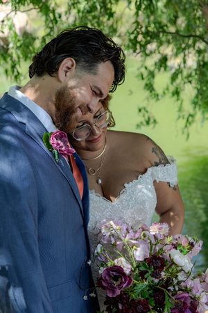 Intimate wedding moments shared under green trees with purple and white floral bouquet details.