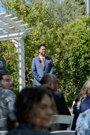 Wedding guests seated outdoors under a white pergola with green foliage in the background.