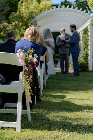 White chairs decorated with floral arrangements line a grassy outdoor wedding ceremony area.