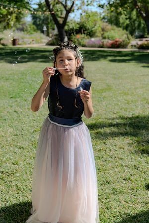 Young flower girl in tulle dress plays with bubbles on grassy lawn.