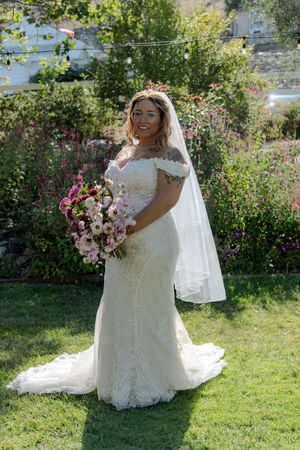 A bride in a lace wedding gown holds a pink and white bouquet while standing on grass near blooming flowers.