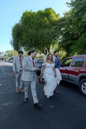 Wedding party walks together in a parking lot wearing gray suits and a white dress on a sunny summer day.