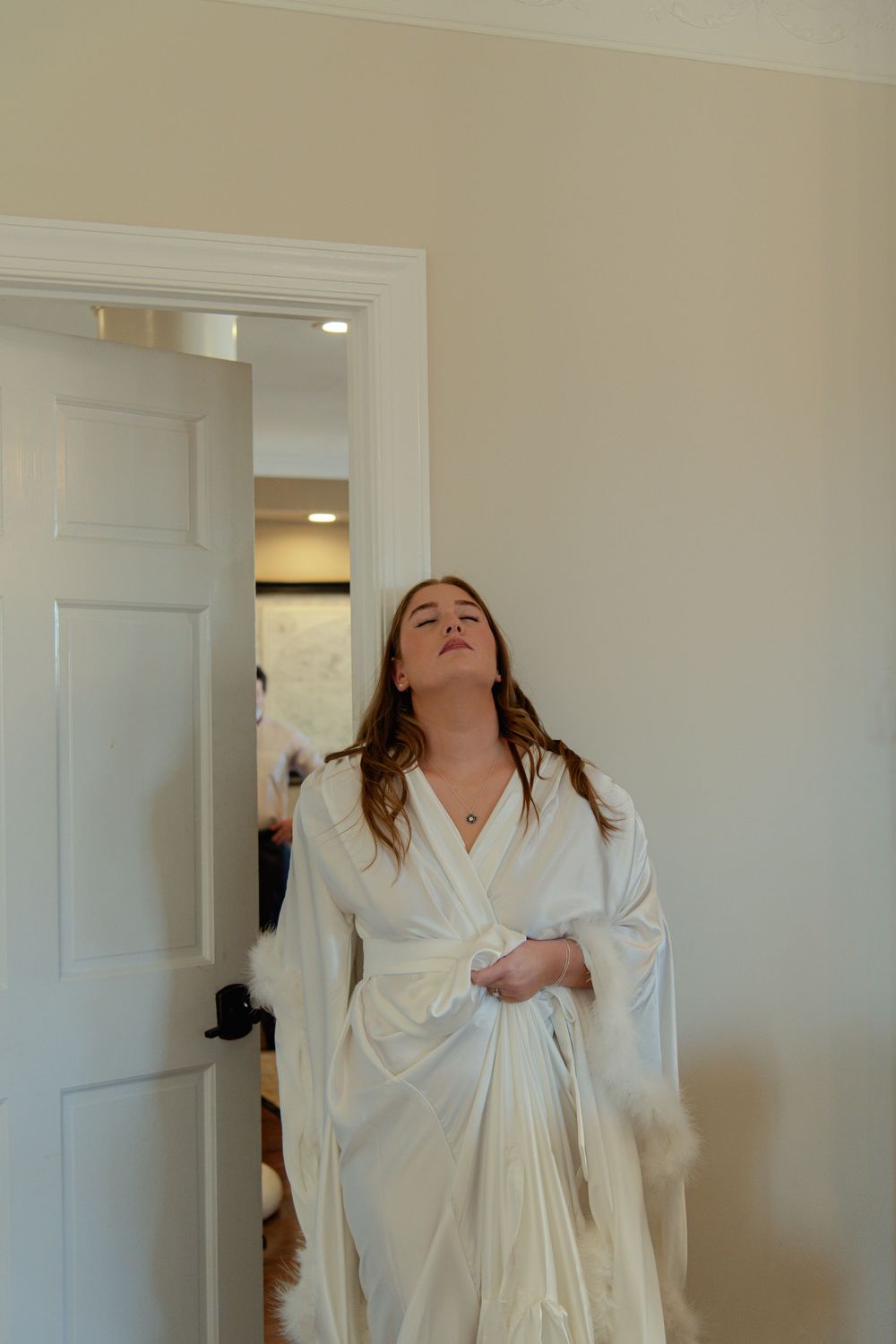 Person wearing a white bathrobe stands in a doorway with arms raised in an expressive pose.