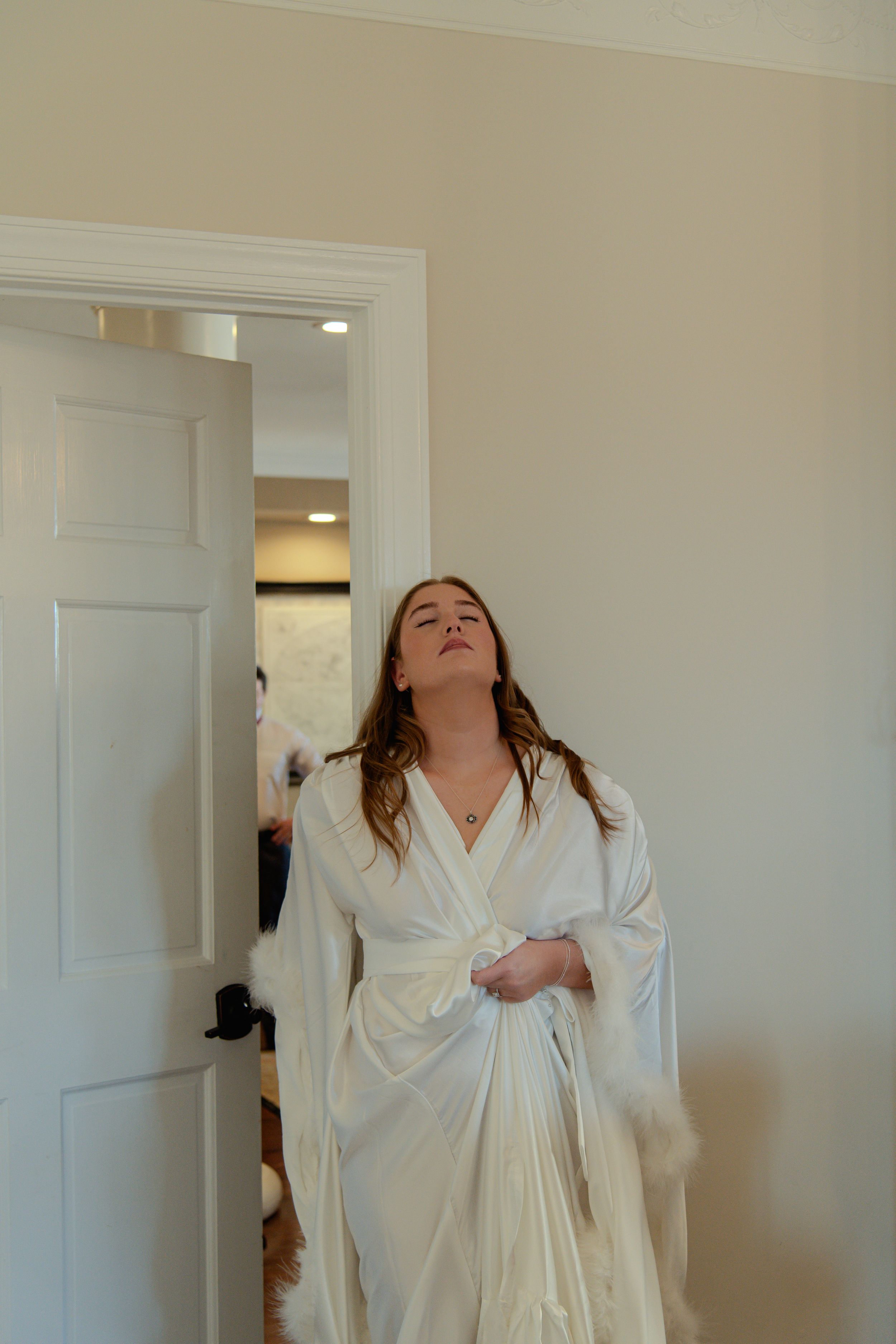 Person wearing a white bathrobe stands in a doorway with arms raised in an expressive pose.