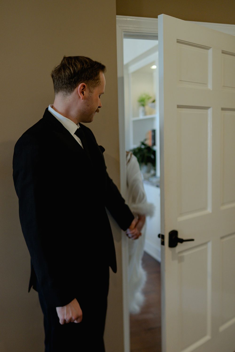 A person in a black suit knocks on a white door in a well-lit hallway.