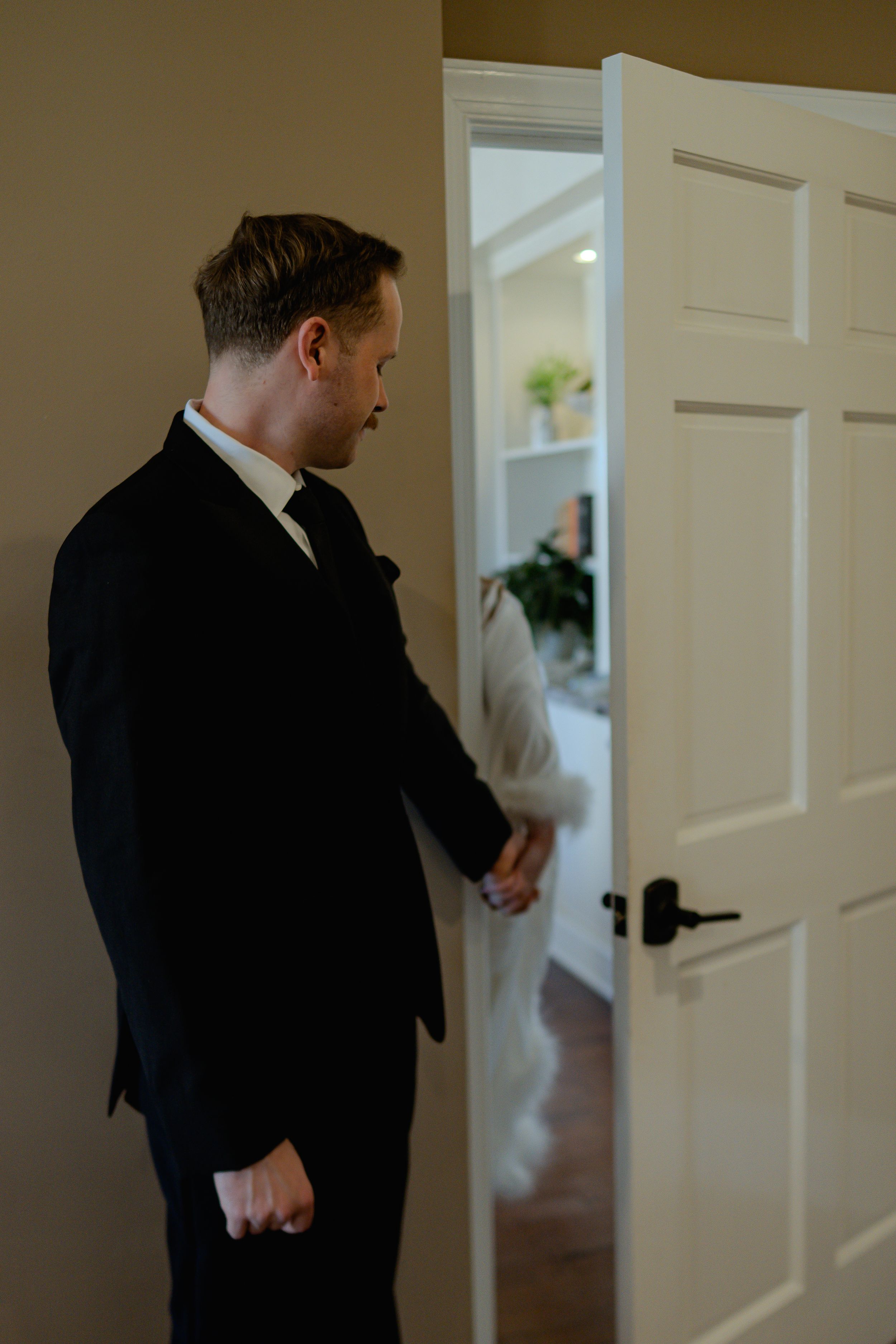 A person in a black suit knocks on a white door in a well-lit hallway.