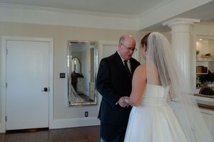 A bride in a white wedding dress gets ready with help in a bright interior room.