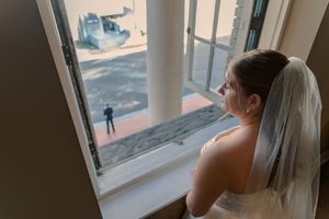 A bride in a white wedding dress looks out a window in a contemplative pose during a wedding day moment.