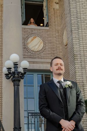 Groomsmen in black suits stand in front of a brick building with ornate lamp posts and architectural details.