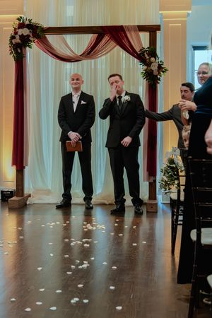 Wedding ceremony setup in an elegant ballroom with white drapes, burgundy accents, and scattered rose petals on dark wood floors.