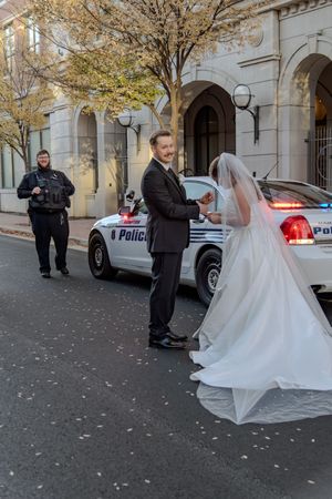 A bride and groom stand near a police car on a city street while an officer watches in the background.