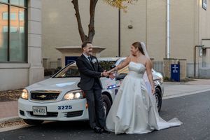 A couple in wedding attire poses with a silver BMW car parked on a city street.