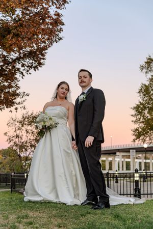 A bride and groom pose outdoors at sunset with autumn trees and a building in the background.