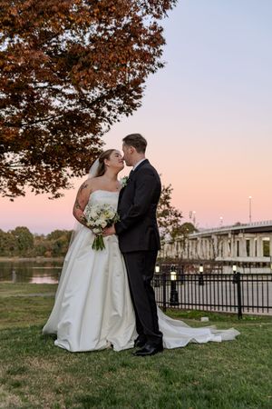 Newlywed couple shares romantic moments at sunset by a waterfront with autumn trees in the background.