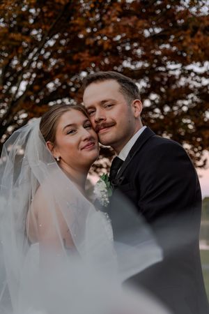 A romantic autumn wedding portrait with a bride in white veil and groom in black suit under orange fall foliage.