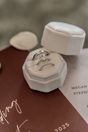 Diamond engagement rings rest on white ring holders with names and dates etched on a brown surface.