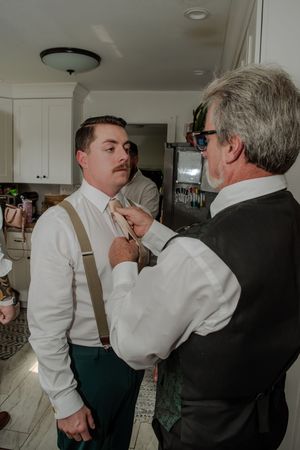 A person helps adjust another's bow tie while getting ready in a kitchen with white cabinets and modern decor.