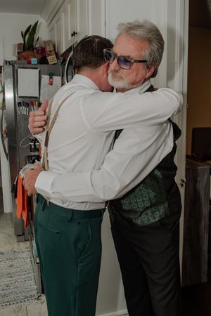 Two people share an emotional embrace in a hallway while wearing contrasting white and dark green outfits.