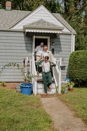 A sequence showing people exiting a small gray house with white trim and walking down a dirt path.