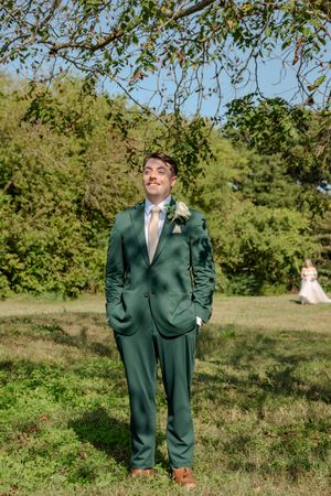 A groom in a dark green suit and brown shoes poses in a sunny outdoor setting with trees in the background.