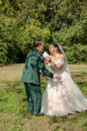 Bride and groom share a joyful dance together on grassy lawn surrounded by trees.