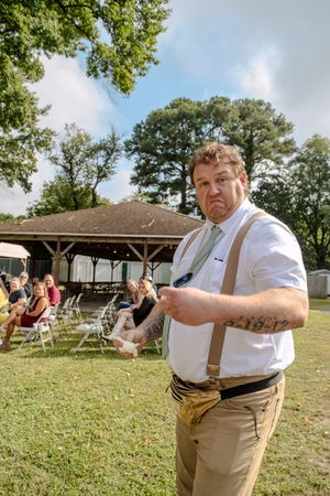 Person in suspenders making expressive gestures during an outdoor wedding ceremony.