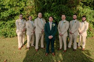 Wedding party members in tan and navy suits pose together on a grassy lawn.