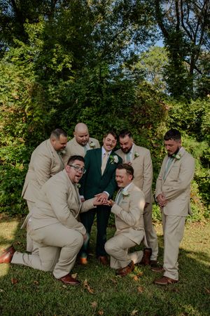 Groomsmen in tan suits and one in green share a playful moment while kneeling on grass during a wedding photoshoot.