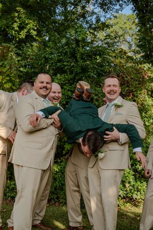 Groomsmen in tan and green suits pose together playfully for wedding photos outdoors.