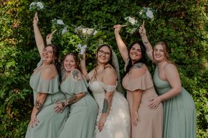 A bridal party in sage green and blush pink dresses pose joyfully with bouquets against a lush garden backdrop.