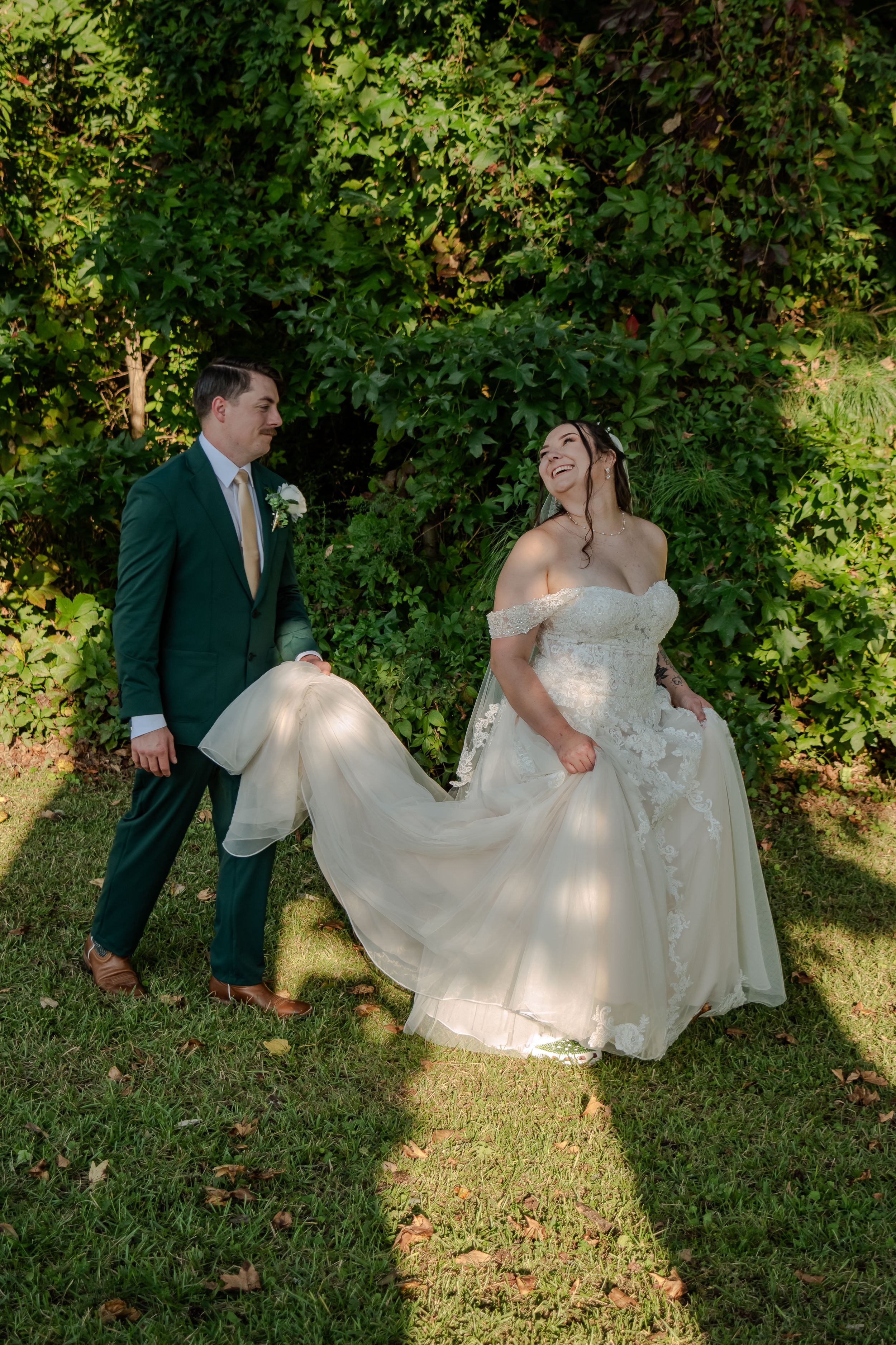 Wedding party poses playfully on grass with flowing white dresses and formal attire in a garden setting.