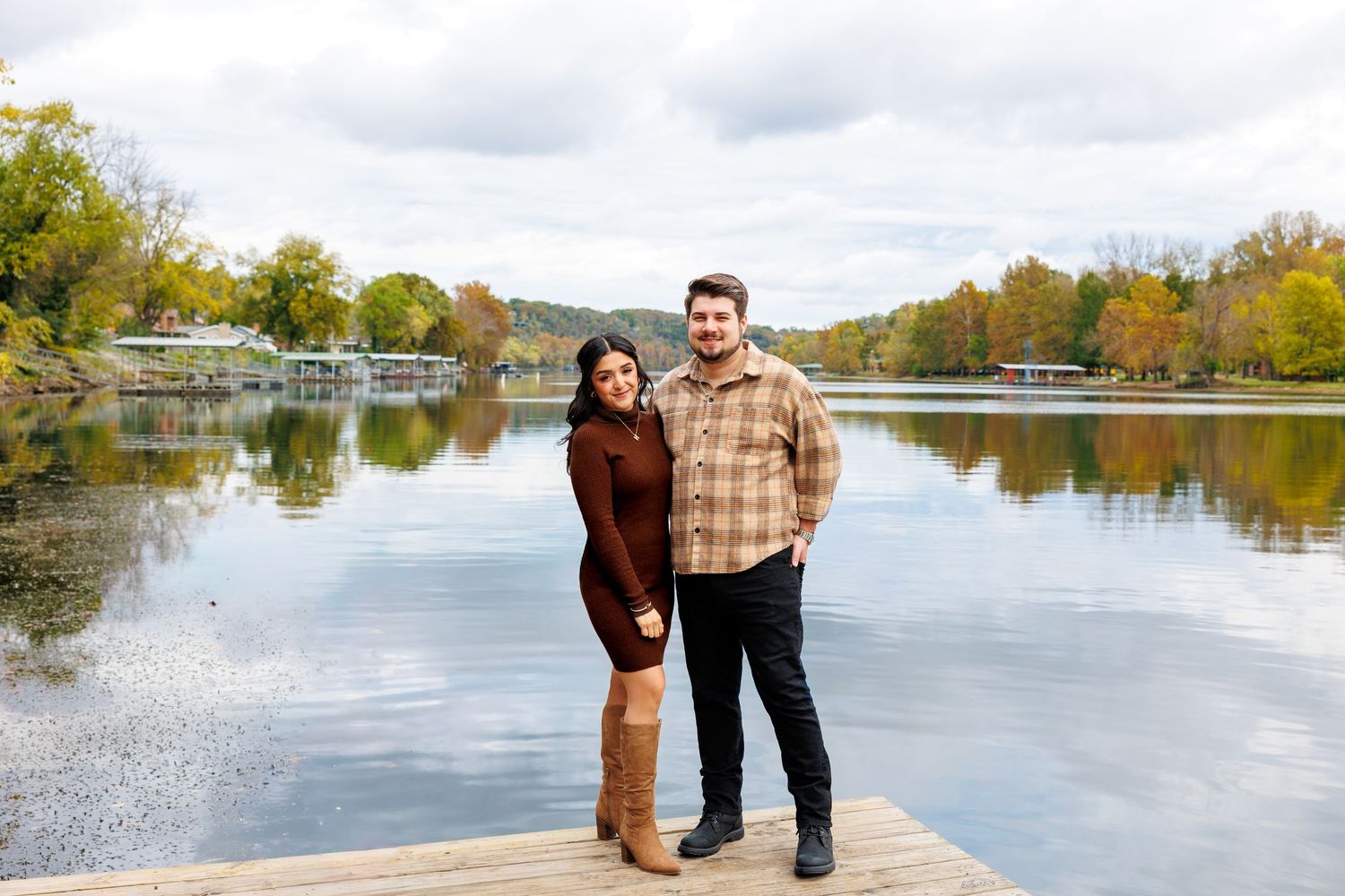 A couple stands together by a serene lake surrounded by autumn trees and mountains in the background.