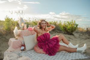 A person in a pink tulle dress and white boots lounges on a beach blanket with pillows at sunset.