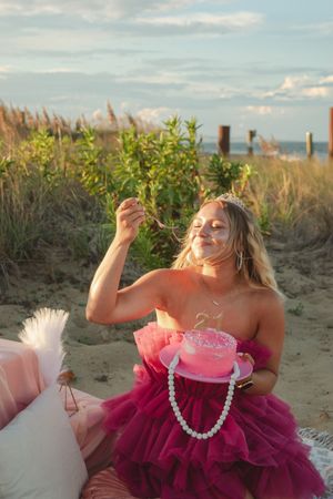 A person in a pink tulle dress poses playfully on a sandy beach at sunset with natural coastal vegetation in the background.