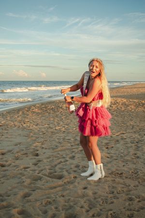 Person in a red dress and white boots dances playfully on a sandy beach at sunset with ocean waves in background.